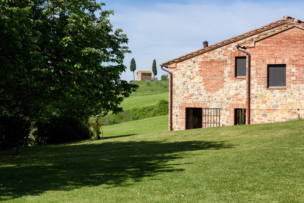 A rustic stone farmhouse is nestled in a verdant landscape. The building features a traditional tiled roof and multiple windows framed by lush greenery. A gently sloping lawn leads toward the rolling hills and distant cypress trees, creating a serene countryside setting.