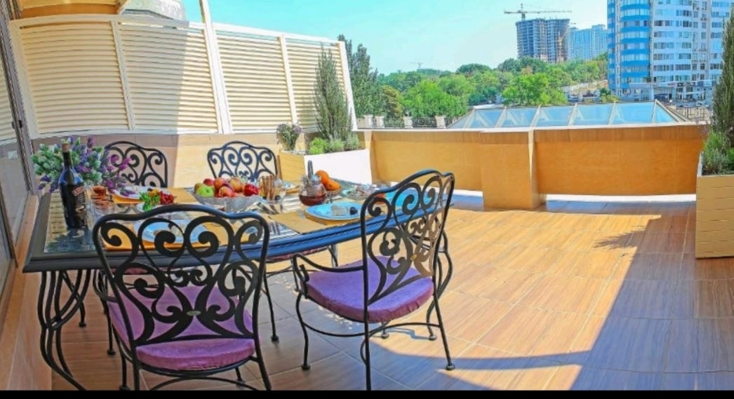 An outdoor terrace is presented with a dining table surrounded by metal chairs featuring decorative backs. A fruit bowl and small plates adorn the table. Lush greenery and modern buildings are visible in the background, under clear blue skies.