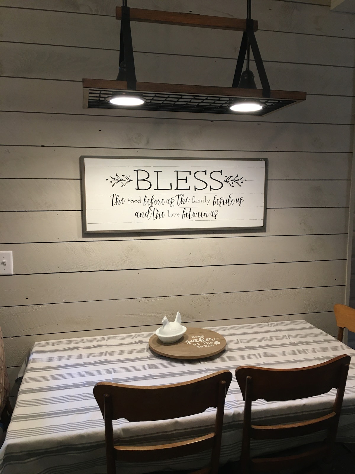 A dining area is highlighted by a wooden table set with neutral-toned linens. Above, a decorative sign with a blessing is displayed on the wall. Two wooden chairs complement the table, enhancing the inviting atmosphere of the space.