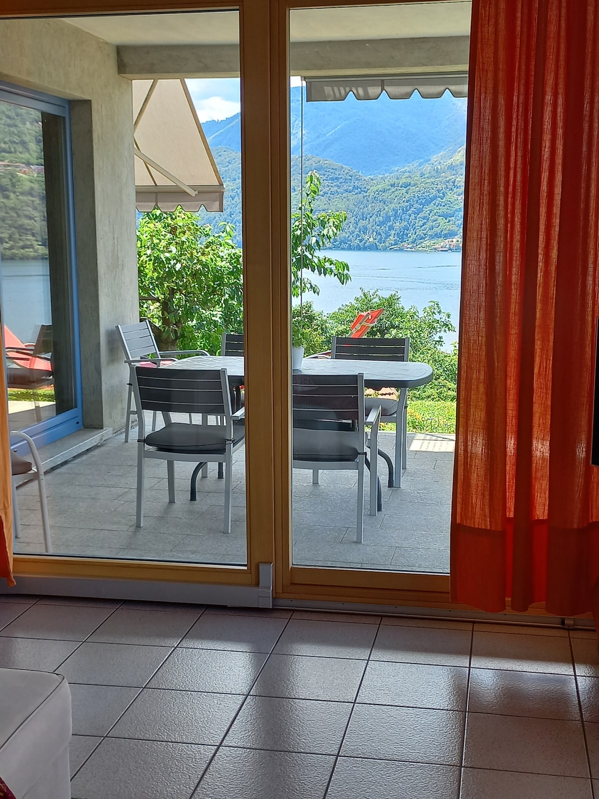 A bright and airy living space transitions to an outdoor patio through large glass doors. A dining table with several chairs is positioned under an awning, surrounded by lush greenery and a view of the lake and mountains beyond.