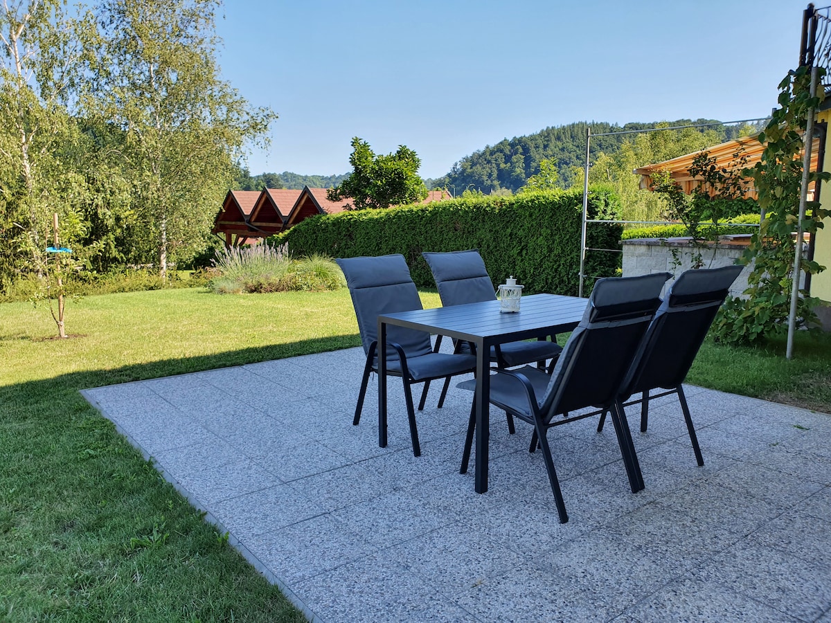 An outdoor dining area features a rectangular table surrounded by six comfortable chairs on a stone-paved patio. Lush green grass and shrubs create a serene garden backdrop, complemented by distant hills and wooden structures, suggesting a peaceful environment for relaxation.