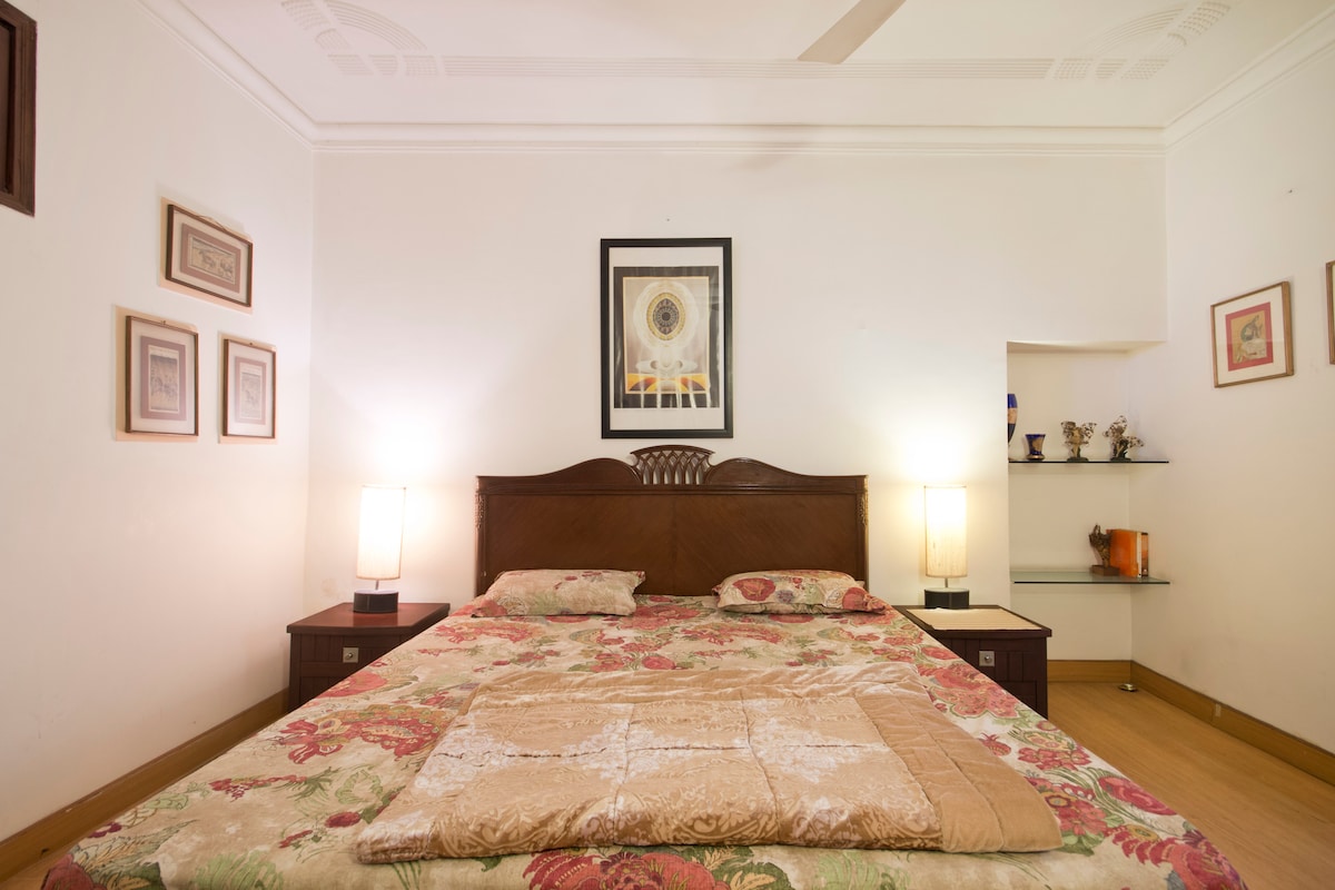A spacious bedroom is centered around a large bed with floral bedding. Two bedside lamps provide soft lighting. Framed artwork hangs on the walls, complementing a small shelf with decorative items. The wooden furnishings add warmth to the serene ambiance.
