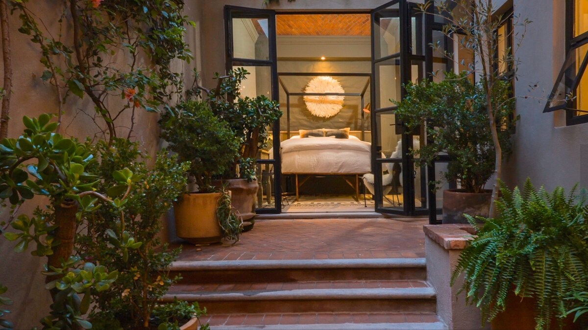 A tranquil courtyard is framed by lush greenery and decorative plants in terracotta pots. Elegant double doors lead to a spacious bedroom, featuring a cozy bed and warm lighting, complemented by a textured ceiling visible in the background.