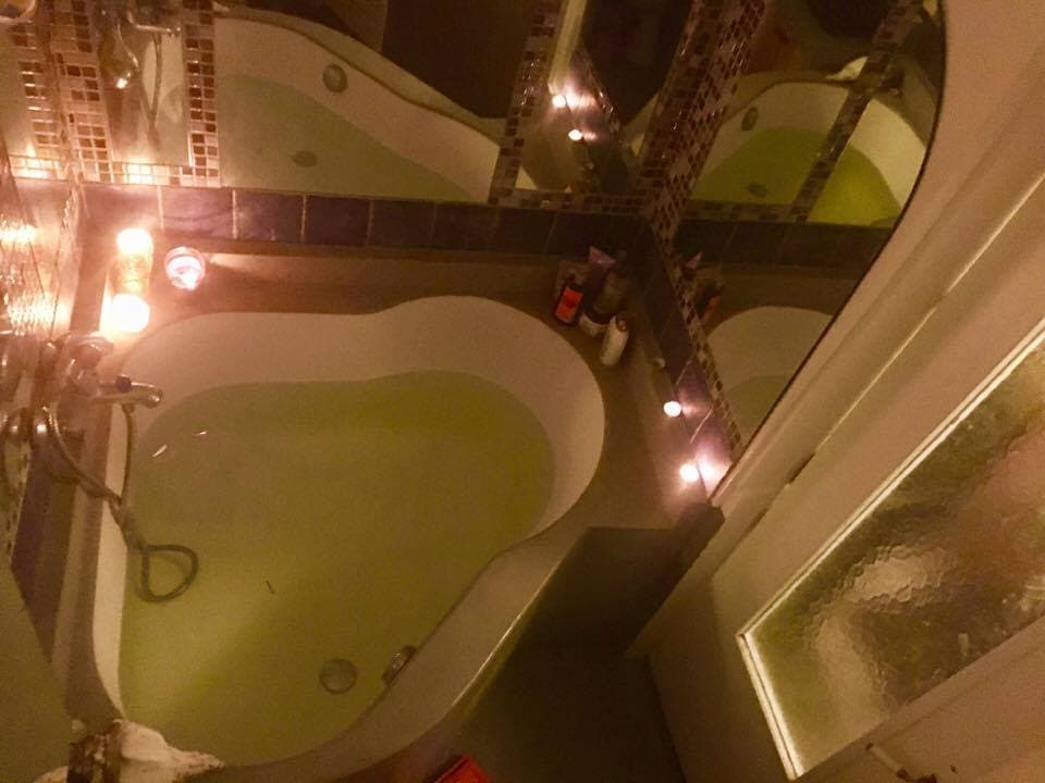 A uniquely shaped bathtub is showcased within a well-lit bathroom, surrounded by reflective tiles and candles that create a calming atmosphere. The gently lit tub water reflects the surrounding light, while various toiletries are neatly arranged nearby.