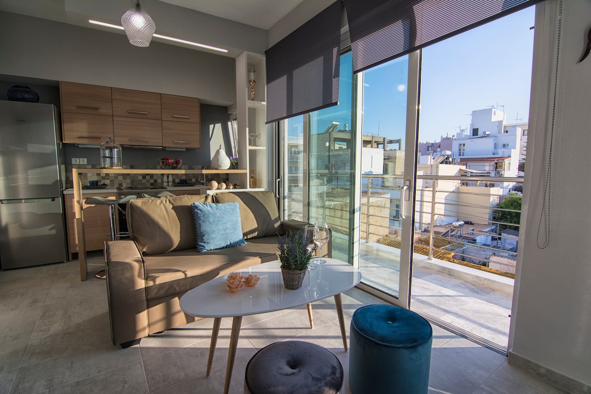 A bright and modern living space is featured, with large glass doors opening to a balcony. A light-colored sofa sits adjacent to a dining table, complemented by a decorative blue cushion and a small plant on the table. Natural light fills the room.