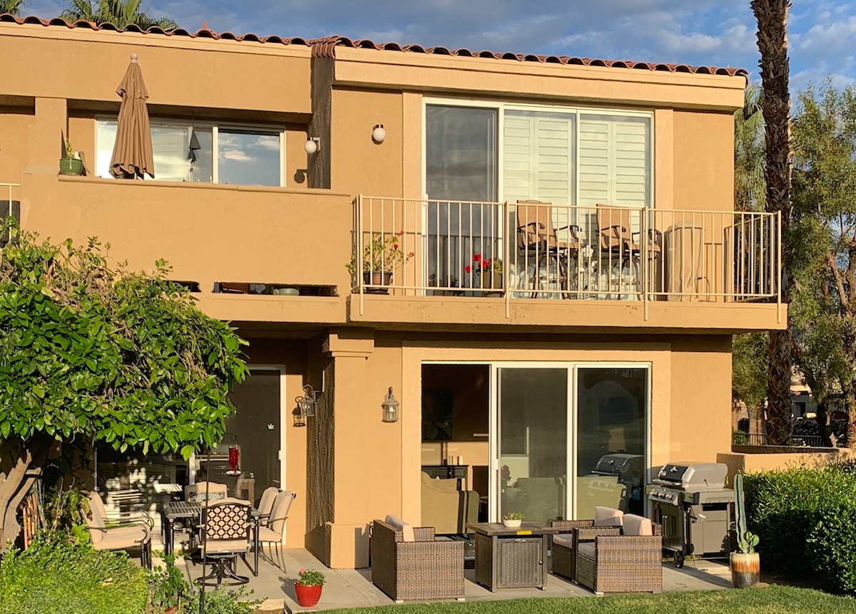 A two-story condo exterior is shown, featuring a spacious balcony with seating for four. A patio area with comfortable furniture is visible below, along with a gas grill. Lush greenery frames the scene, showcasing a welcoming outdoor space surrounded by a serene environment.