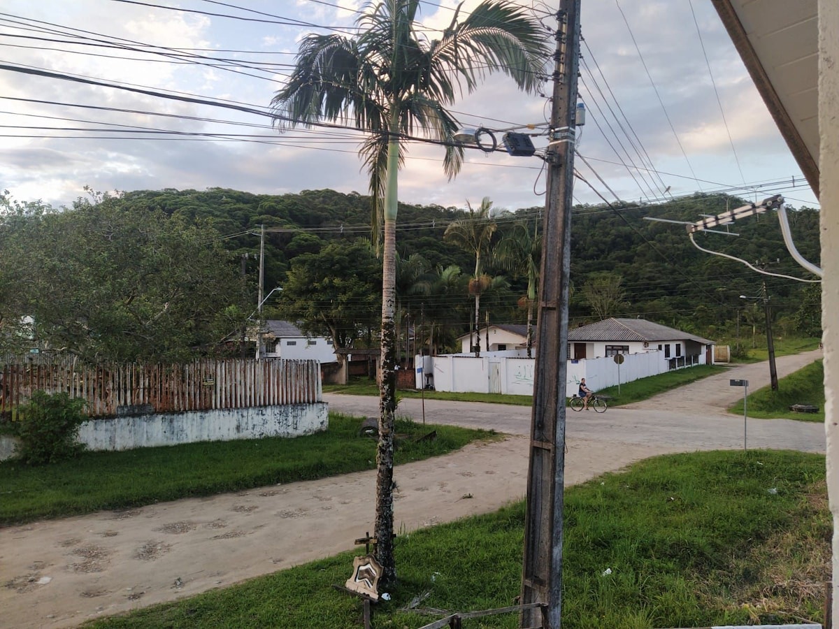 A view of a residential area is presented, featuring a winding dirt road bordered by lush greenery. On the right, a cyclist is seen near the intersection. Power lines and palm trees are visible, framing the tranquil environment of the surrounding hills.