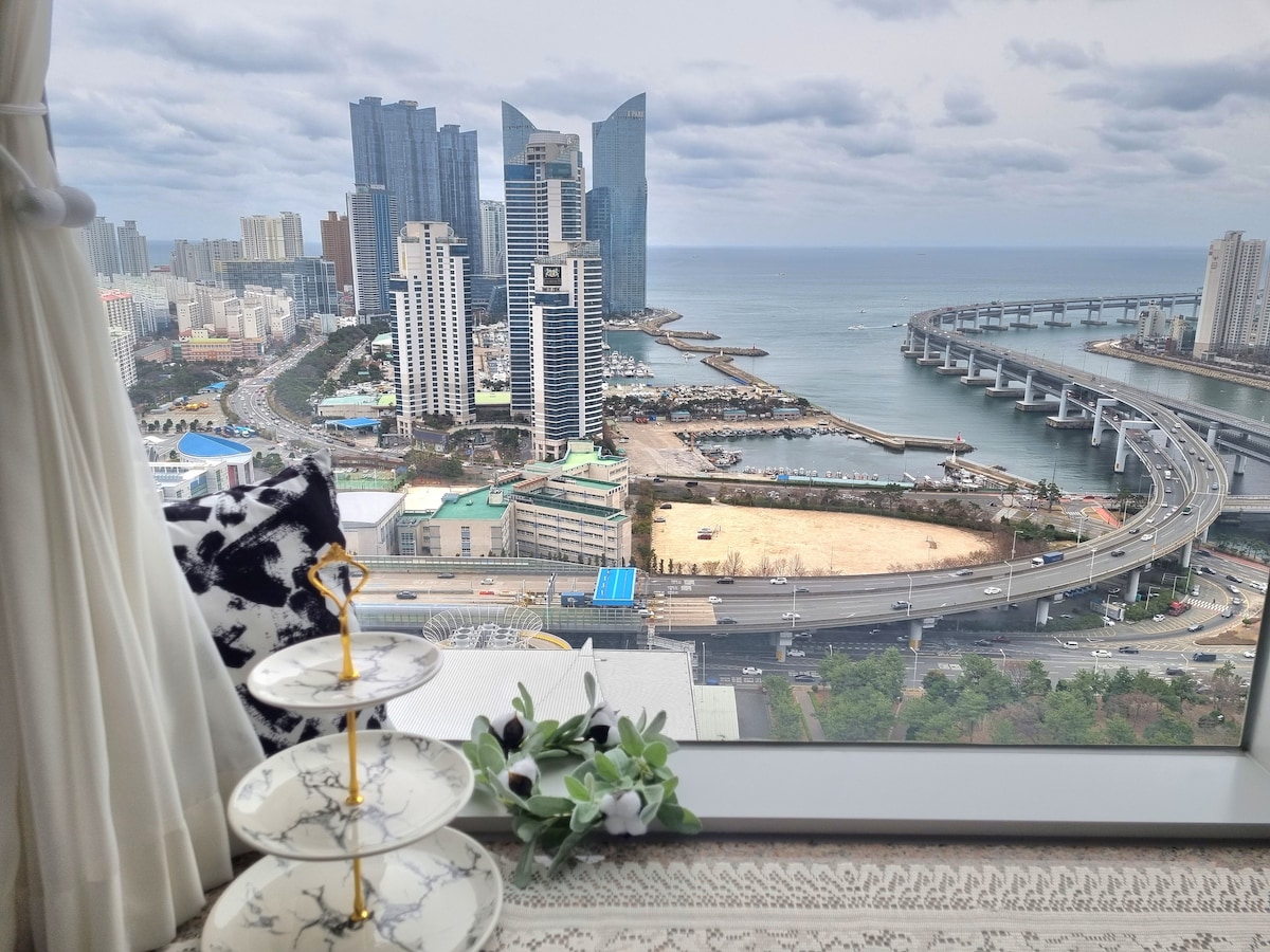 A panoramic view showcases the coastal city, featuring modern skyscrapers and the bridge over the water. A three-tiered stand decorated with minimalistic floral accents is placed near the window, inviting guests to enjoy the scenery while relaxing indoors.