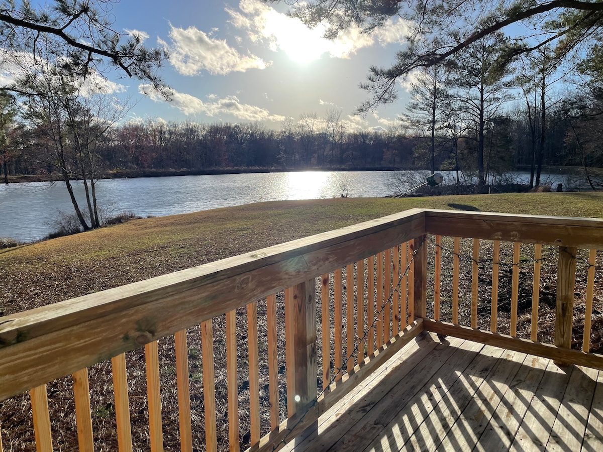 A wooden deck overlooks a tranquil lake, reflecting sunlight on the water's surface. Lush greenery surrounds the area, with trees lining the water's edge and a clear sky in the background. The space is designed for relaxation and enjoying scenic views.