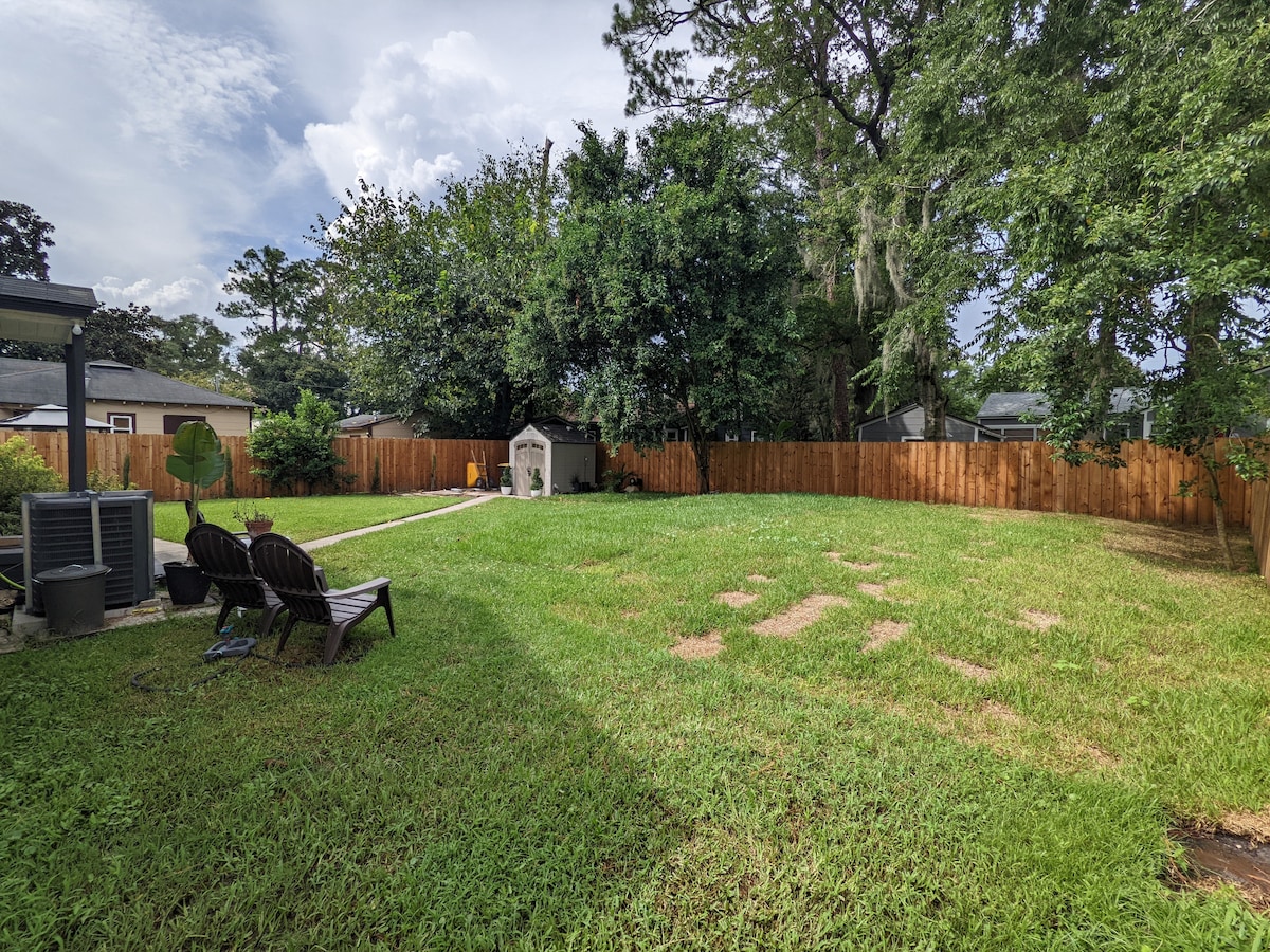 A spacious fenced backyard is shown, featuring a neatly maintained lawn surrounded by tall trees and lush greenery. Two wooden lounge chairs are positioned on one side, facing a small storage shed in the rear. A pathway leads through the grass, creating a serene outdoor setup.