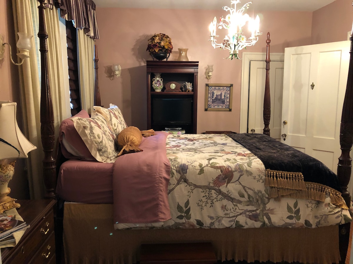 A classic bedroom features a four-poster bed dressed in floral bedding with coordinating accent pillows. A dark throw blanket is draped over the foot of the bed. Subtle pink walls are complemented by elegant decor, including a chandelier and a bedside lamp, creating a cozy ambiance.