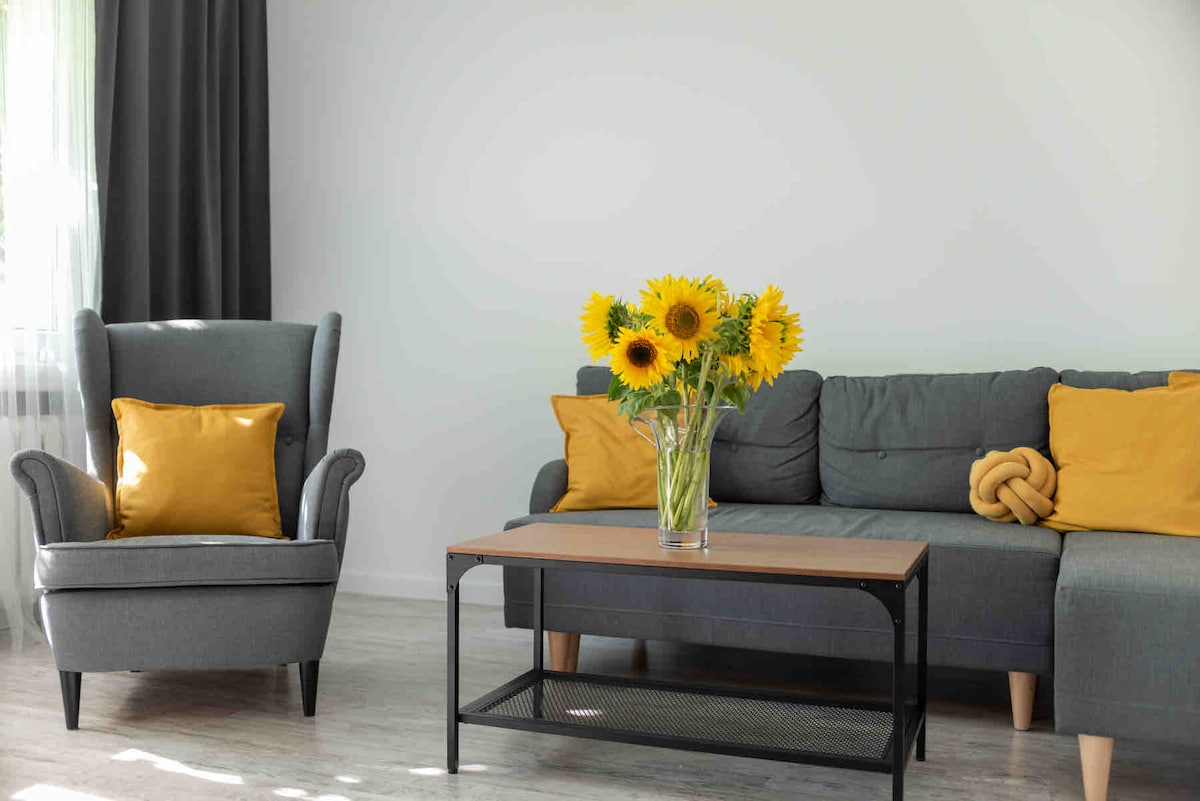 A comfortable living area is presented, featuring a gray sofa adorned with yellow cushions. A simple coffee table sits at the center, supporting a vase filled with fresh sunflowers. A light curtain gently filters natural light, enhancing the serene ambiance of the space.