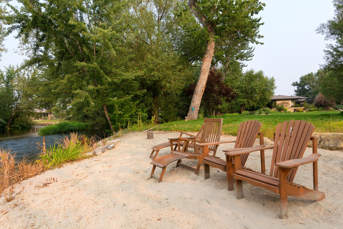 Three wooden Adirondack chairs are positioned on a sandy area beside the river, surrounded by greenery. Tall trees provide shade, and a variety of plants and grass are visible in the background, creating a serene atmosphere by the water.