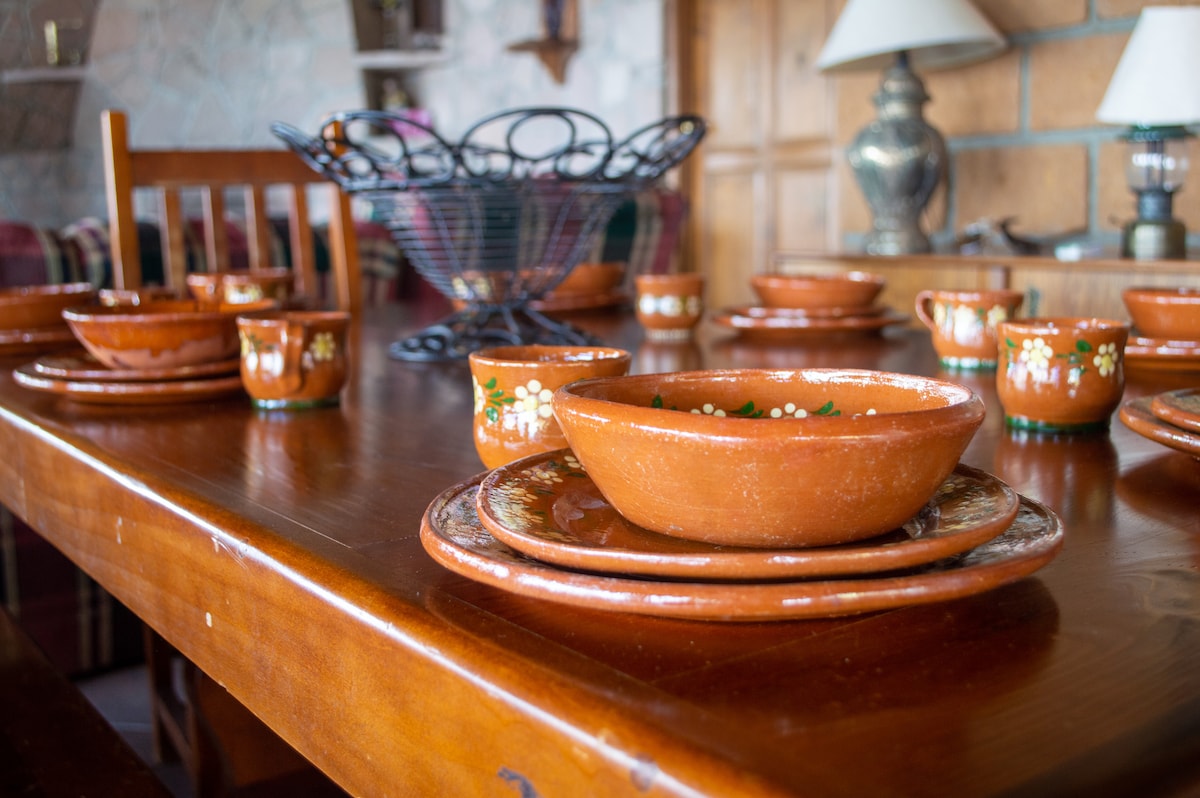 A rustic wooden dining table is set with handcrafted clay dishes, including various bowls and plates. The table is adorned with decorative cups and a wire fruit basket. The inviting layout suggests a space for shared meals and gatherings.