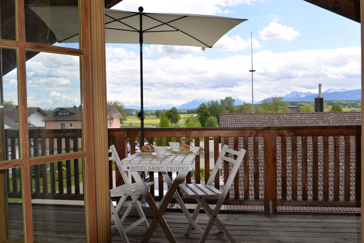 A balcony features a small table set for outdoor dining, complete with pastries and drinks. A large umbrella provides shade. The view extends to rolling greenery and distant mountains under a partly cloudy sky.