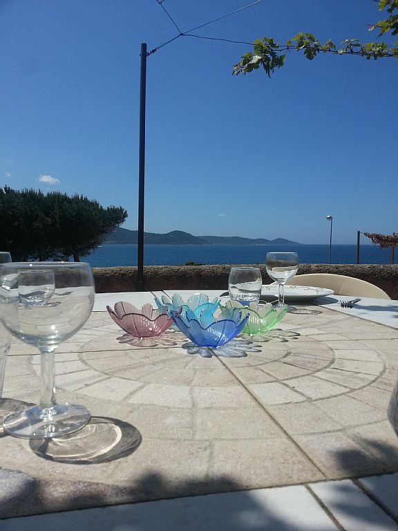A dining table set outdoors displays colorful glass dishes and wine glasses. The view of the sea is visible in the background, framed by trees and distant hills. The clear sky enhances the overall ambiance of the setting.