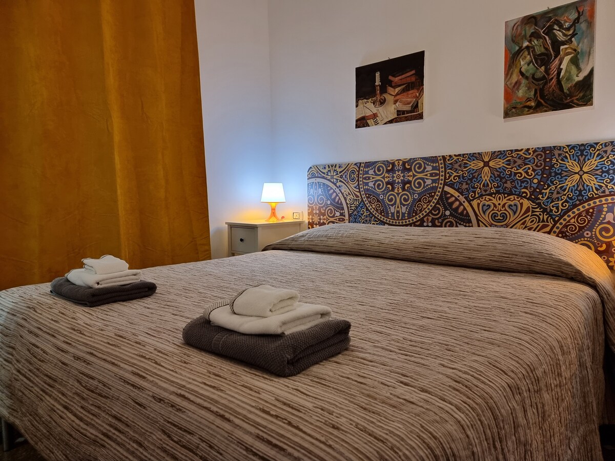 A spacious bed is placed against a patterned headboard, with neatly arranged towels on top. A bedside lamp emits a soft glow, complementing the warm yellow curtain that drapes beside the window. Two art pieces hang on the wall, adding a touch of character.