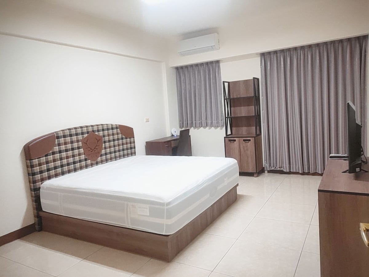 A spacious bedroom is presented with a large bed featuring a plaid headboard. A minimalist desk and chair are positioned by a window, while a tall bookshelf stands nearby. Light-colored tiles and soft curtains enhance the overall brightness of the room.