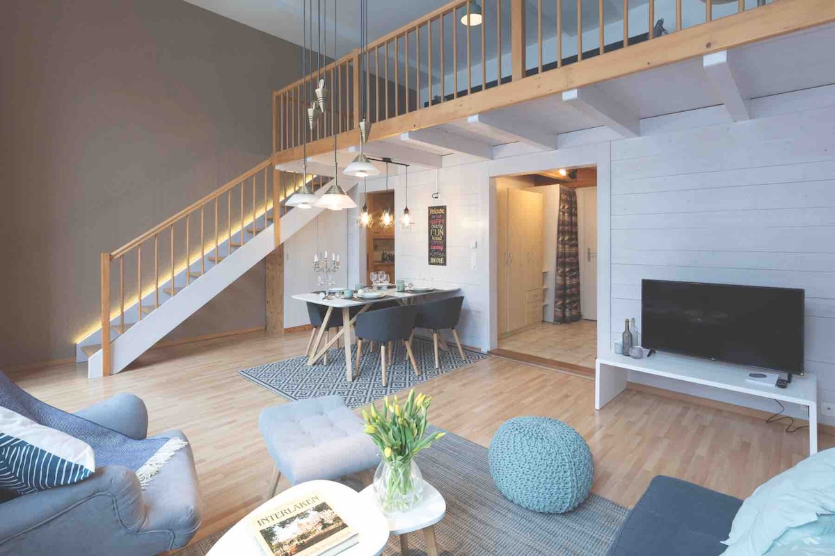 An open living area features a cozy seating arrangement with a soft blue armchair and a light-colored sofa. A dining table is set for guests, and an exposed staircase leads to the upper gallery. Natural light fills the space from large windows.