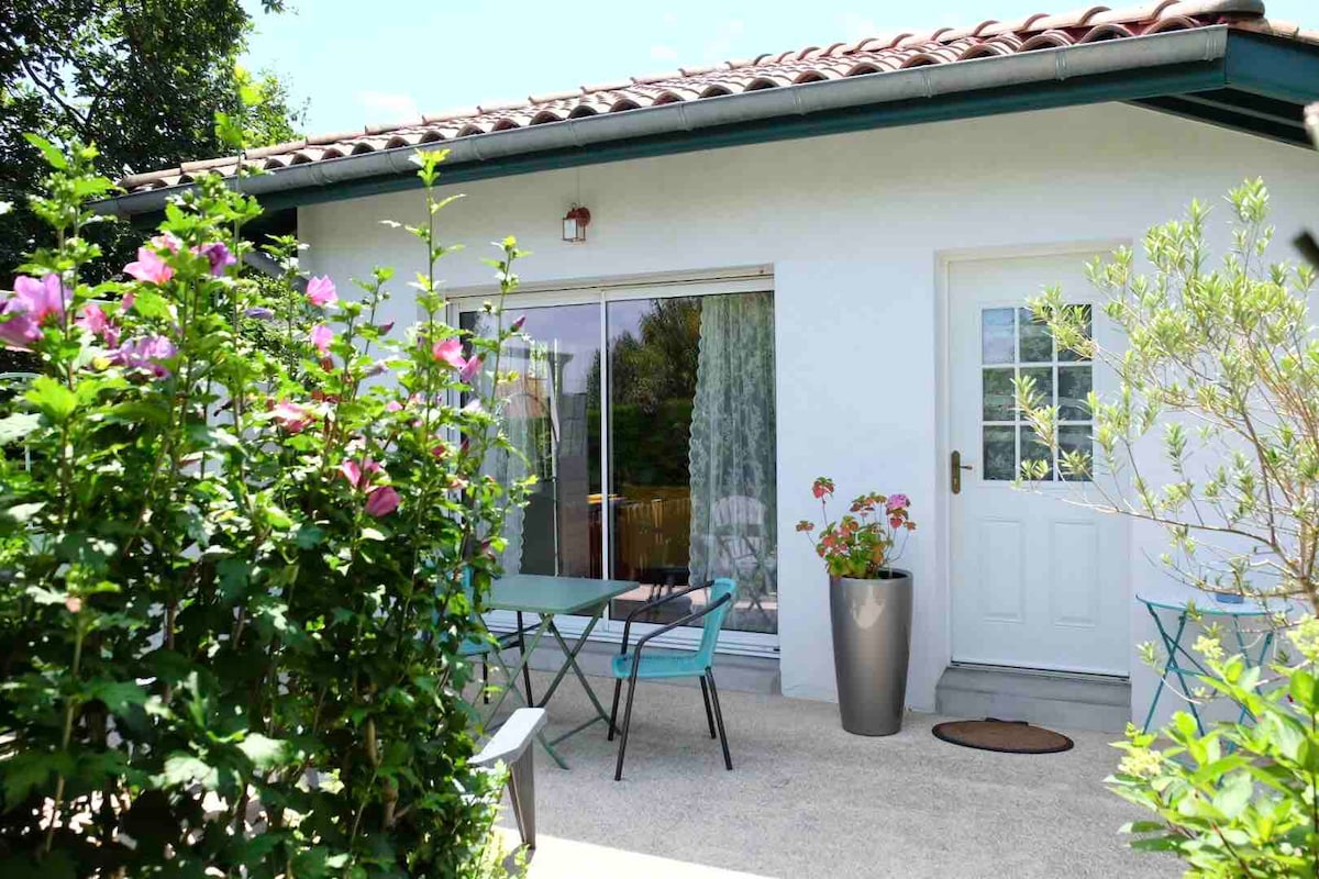 The exterior of the charming house is framed by vibrant flowers and greenery. A small table with chairs is positioned on a patio area, visible through glass doors that invite natural light into the space. A welcoming entrance is outlined by a potted plant.