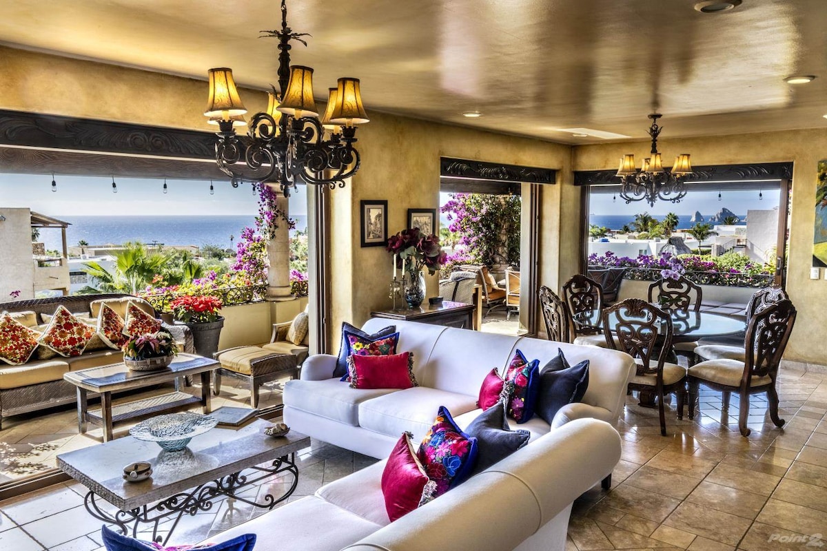 A spacious living area features large windows showcasing ocean views. Comfortable seating is arranged around a coffee table, adorned with decorative cushions. A dining area with elegant furniture is visible, enhanced by vibrant floral accents from nearby plants.