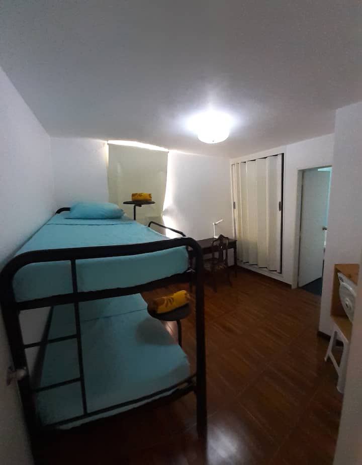 Izba N 2: Výhľad na poschodovú posteľ, s písacím stolom, stoličkou, ventilátorom, šatníkom na dverách na akordeóne 