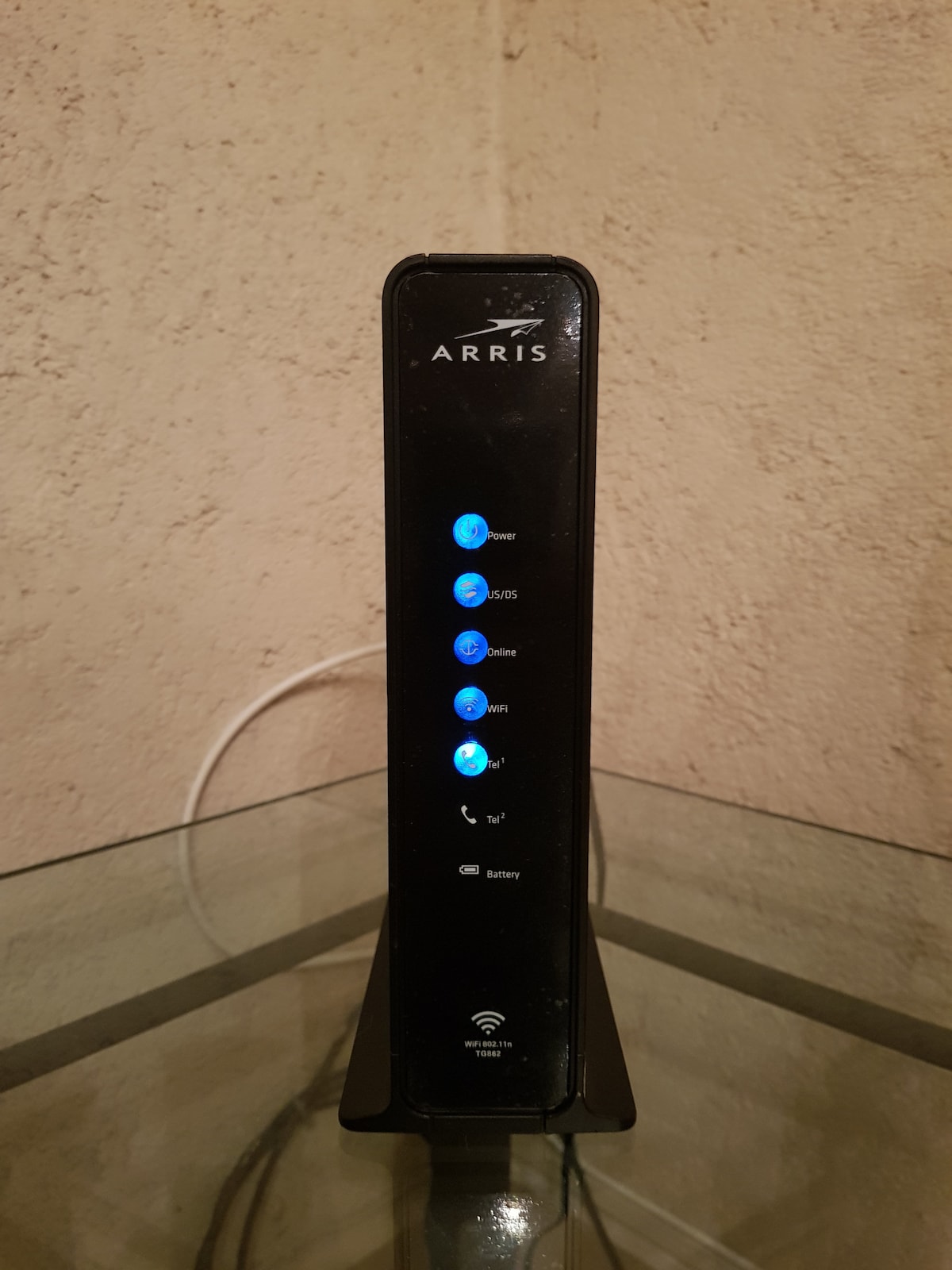 A black internet modem is positioned on a glass surface. It features multiple illuminated indicators, displaying power, connectivity status, and Wi-Fi availability. The modem's sleek design is complemented by a neutral background, enhancing visibility.