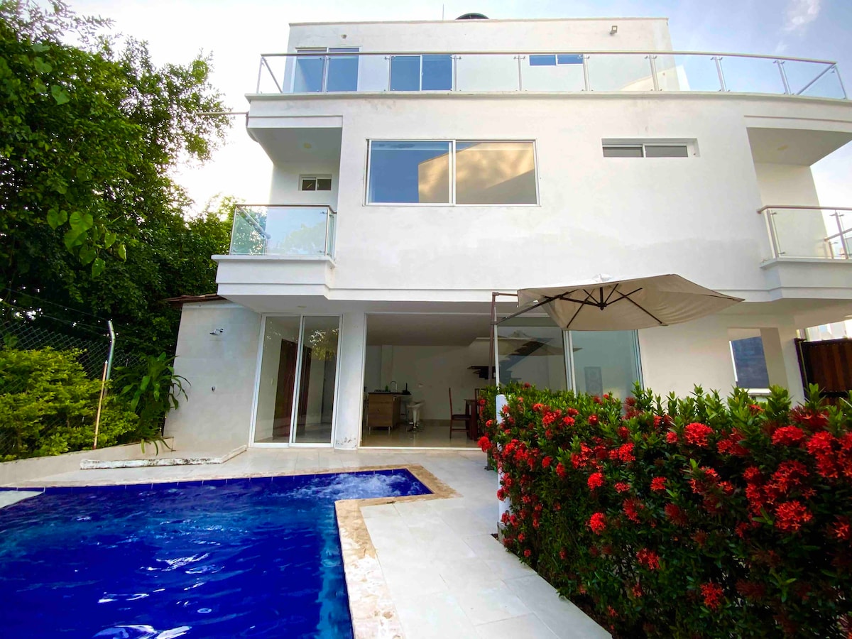 The exterior of a modern structure features large windows and sliding glass doors, allowing natural light to fill the interior. A private pool, bordered by a stone deck, is accompanied by a shaded seating area under an umbrella. Vibrant flowering shrubs enhance the tropical atmosphere.