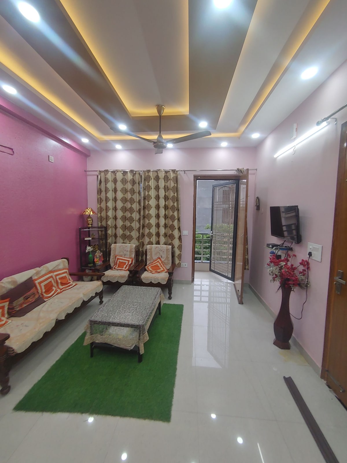 A vibrant living area features comfortable seating arrangements, including a sofa and chairs with decorative cushions. A centerpiece table is positioned on a green rug. Soft lighting highlights the room's décor, while a door opens to an outdoor view. A mounted TV is also present.