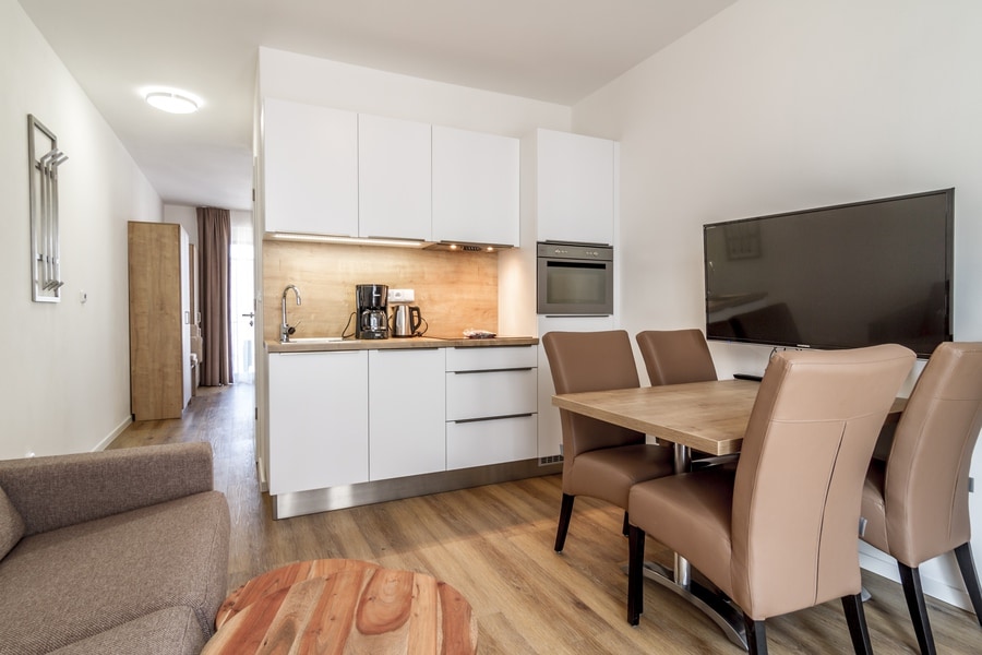 A modern kitchen area features sleek white cabinetry and a wooden dining table with four cushioned chairs. Stainless steel appliances, including a coffee maker and microwave, are visible. A cozy living space is indicated by a soft brown sofa, while a large flat-screen television is mounted on the wall.