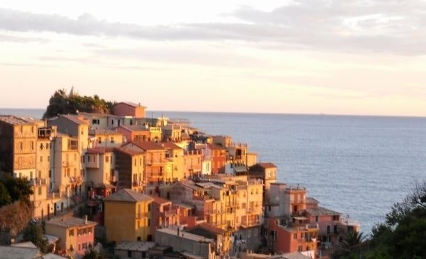 A coastal view of Manarola captures colorful houses bathed in warm sunset light, arranged in a cascading manner on rugged cliffs. The serene sea stretches into the horizon, with soft waves reflecting the evening sky's hues.