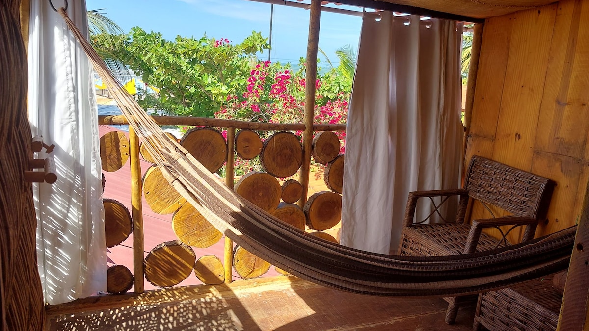 A relaxing outdoor area features a comfortably hung hammock and a woven chair, surrounded by wooden accents. Light filters through white curtains, and vibrant greenery with pink flowers enhances the inviting view.
