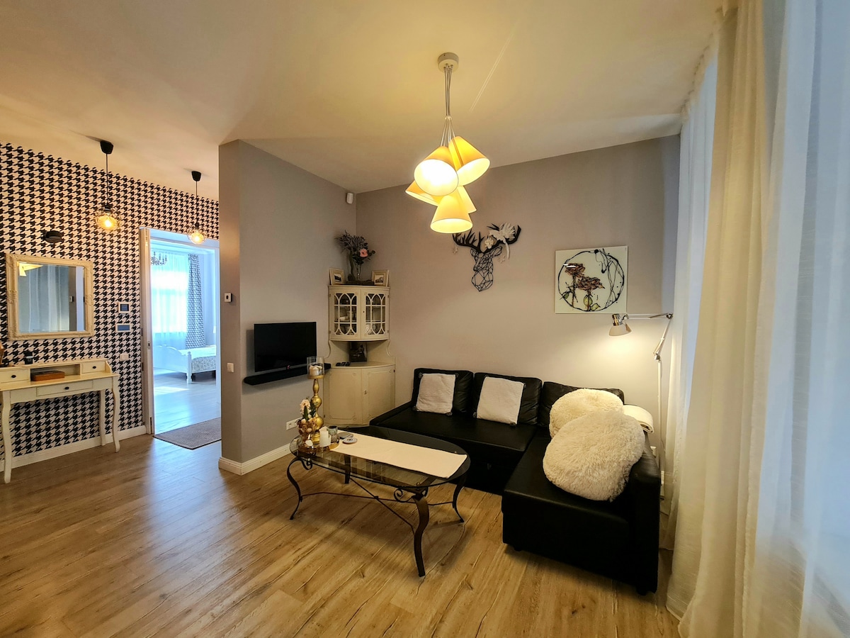 A modern living area features a black sectional sofa adorned with plush white cushions. A sleek coffee table occupies the center, complemented by a decorative lamp and wall art. Light is filtered through sheer curtains, enhancing the cozy ambiance of the space.