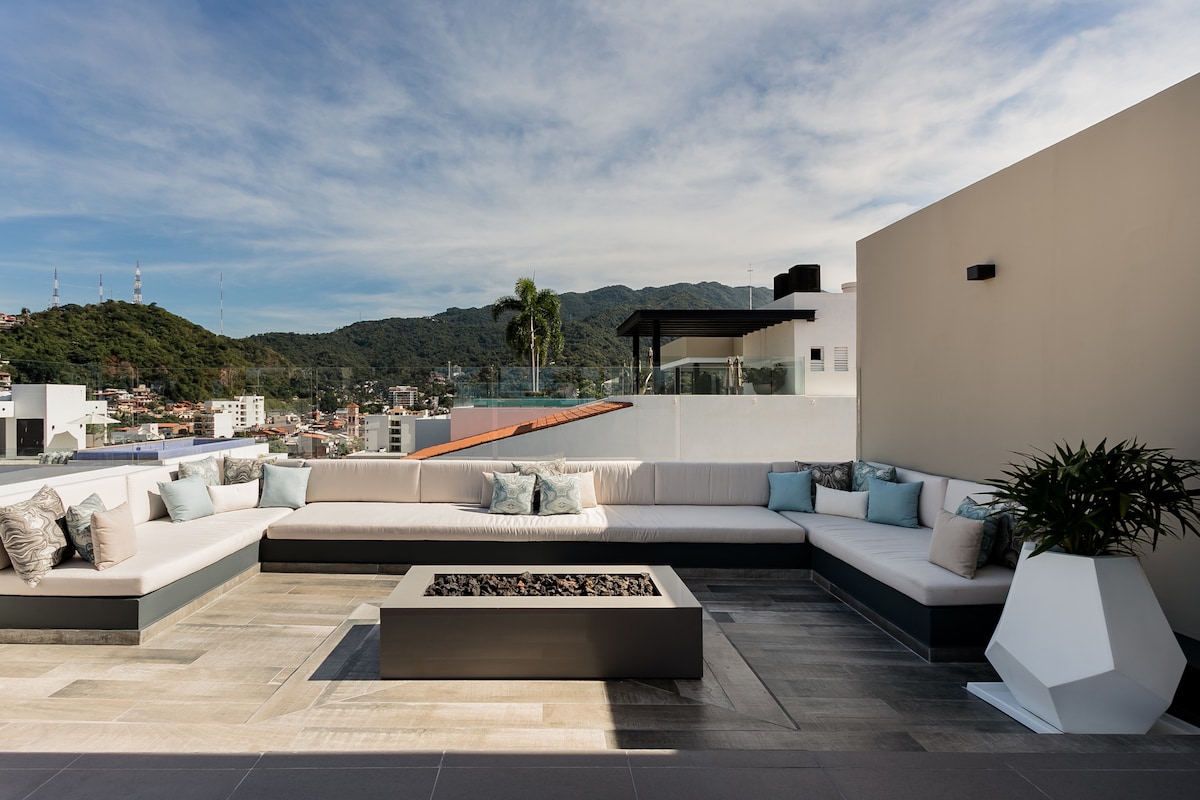 A spacious rooftop lounge area features ample sectional seating adorned with decorative pillows. A modern fire pit is centrally positioned on the sleek tiled floor, surrounded by scenic views of the mountains and cityscape in the background.