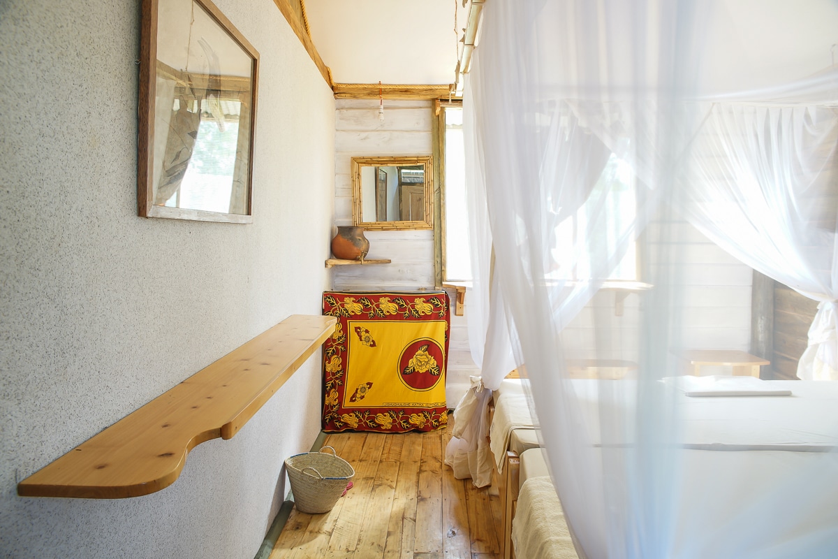 The airy interior features wooden flooring and walls, complemented by soft natural light filtering through large windows adorned with mosquito nets. A handcrafted wooden wash basin is positioned beside a colorfully draped bench, enhancing the room's earthy aesthetic.