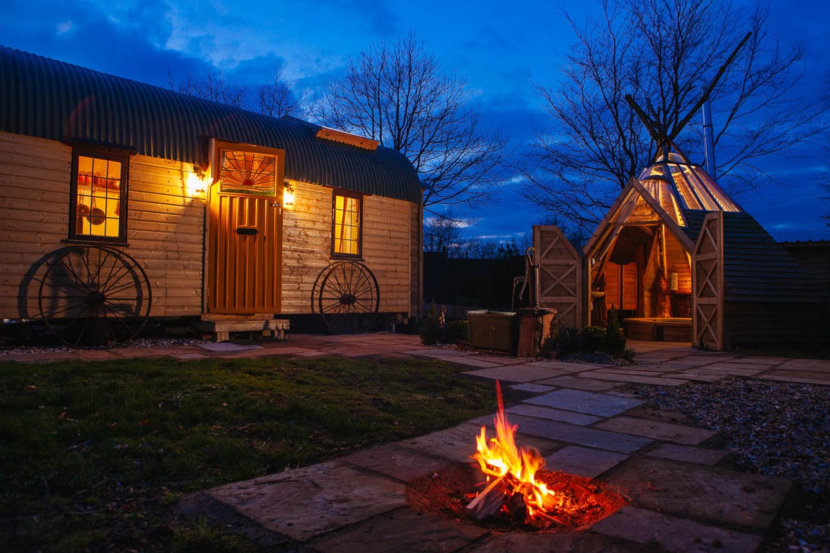 A rustic wagon and wigwam are illuminated as twilight descends. A warm fire pit flickers in the foreground, surrounded by neatly laid stone paving. The wooden wagon features a welcoming door, while the wigwam, topped with a conical roof, offers a glimpse of cozy gathering space.