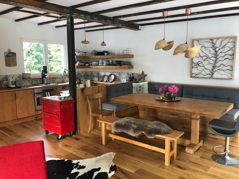 The kitchen area features wooden cabinetry with an open shelf displaying dishware. A large wooden dining table with a bench is central to the space, accompanied by modern pendant lights above. A red rolling kitchen cart adds a pop of color, while warm wood flooring completes the inviting atmosphere.