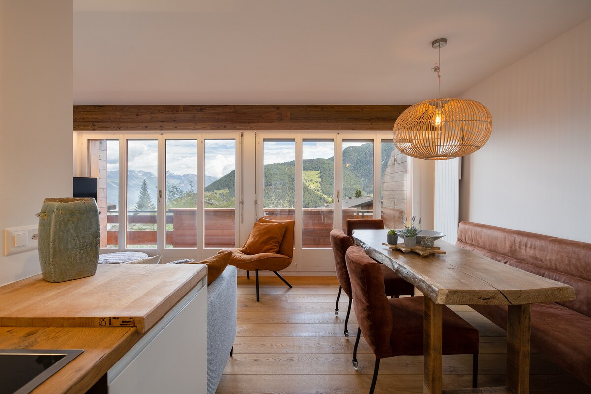 A bright living area features large glass doors opening to a balcony with mountain views. A wooden dining table accommodates six, surrounded by upholstered chairs. Cozy seating is arranged nearby, complemented by natural wood elements and a pendant light above.