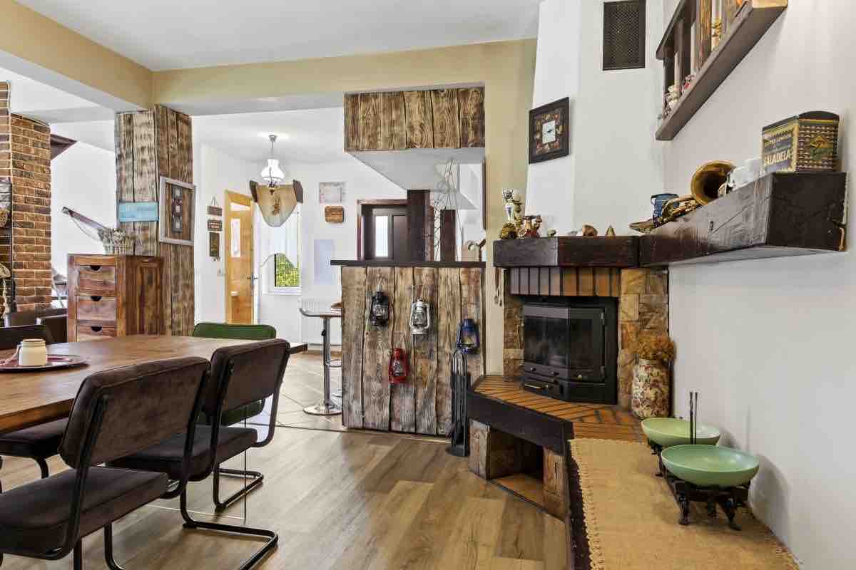 A rustic dining area features a table with six chairs, complemented by an exposed brick wall. A cozy fireplace is set against a wooden accent wall. Decorative items are neatly arranged on shelves, adding character to the space, with natural light flooding in from the adjacent hallway.