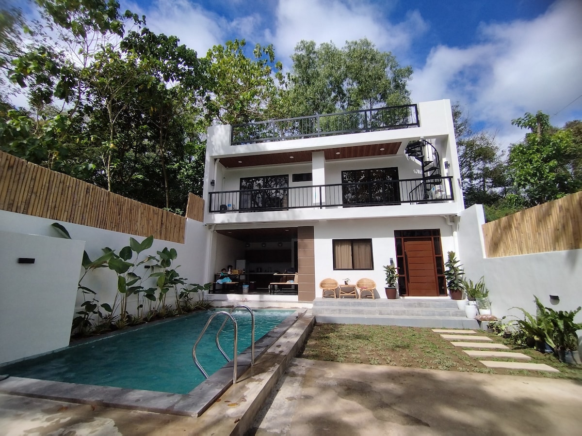 A modern two-story home features a private pool surrounded by greenery. The outdoor area includes a spacious patio with lounge chairs and a covered seating space, inviting relaxation under the trees. Large windows provide a view of the natural surroundings.