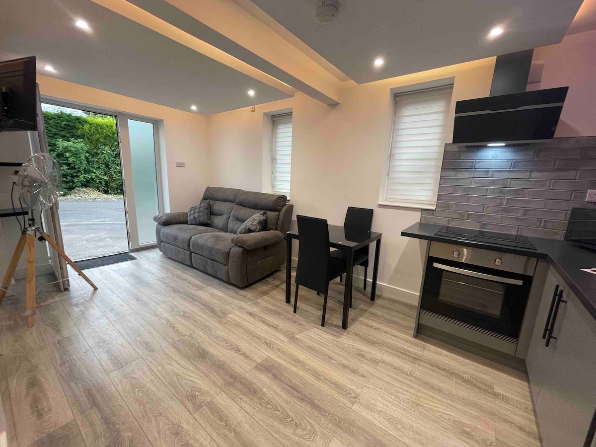 A compact living area features a comfortable gray sofa and a small dining table with black chairs. Natural light filters in through sliding glass doors, which lead to a small outdoor space. A modern kitchen area with sleek cabinetry is visible in the background.