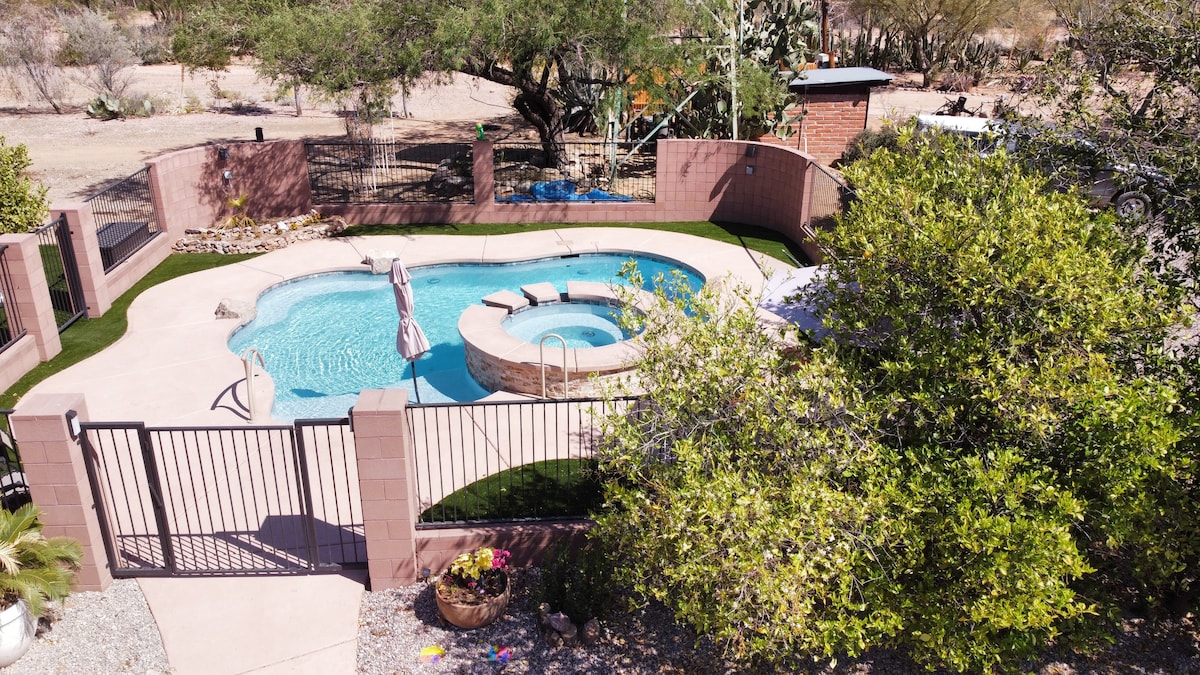 A fenced outdoor area features a serene pool with a natural stone hot tub beside it. The pool is surrounded by artificial grass and desert landscaping, with a few shaded lounge chairs nearby, creating a tranquil space for relaxation.