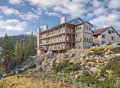 A multi-story building is positioned on a rocky terrain, surrounded by lush greenery and coniferous trees. Large balconies are visible on the upper floors, offering views of the landscape. The sky is partly cloudy, and the structure exhibits a blend of architectural styles.