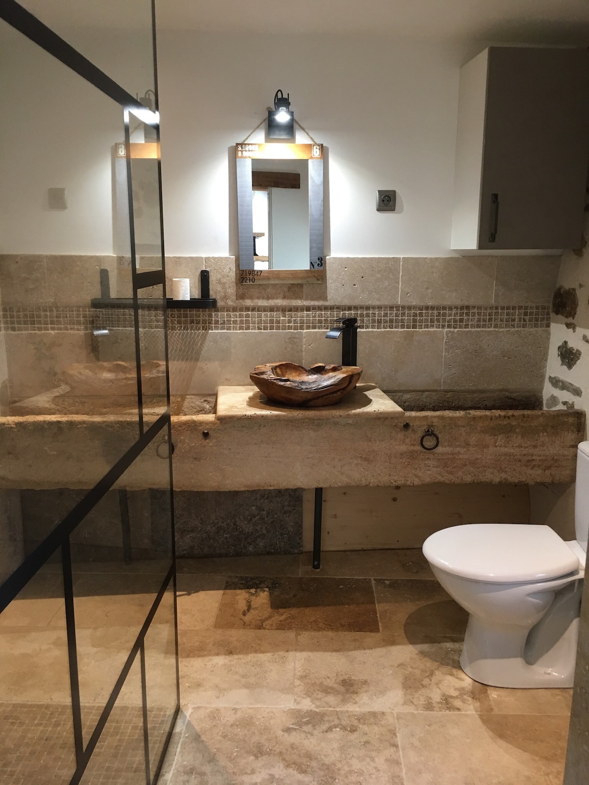 The bathroom features a modern design with natural stone accents. A large glass shower is visible, along with a sleek vanity made of stone. A wooden bowl serves as the sink, while subtle lighting highlights the space. A white toilet complements the overall aesthetic.