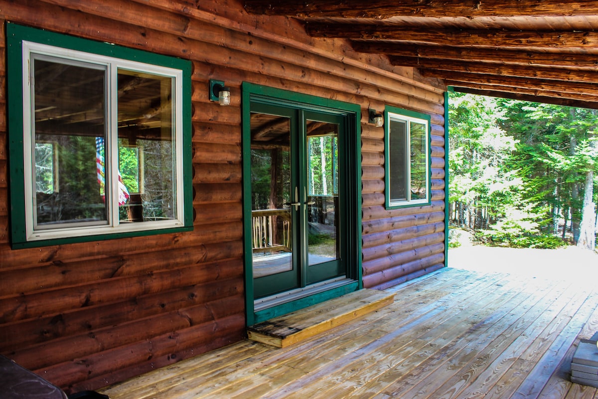 The entrance to the cabin features a wooden deck with a slightly raised step leading to the double doors. Two windows are present, providing natural light, with views of the surrounding woods evident through the glass.