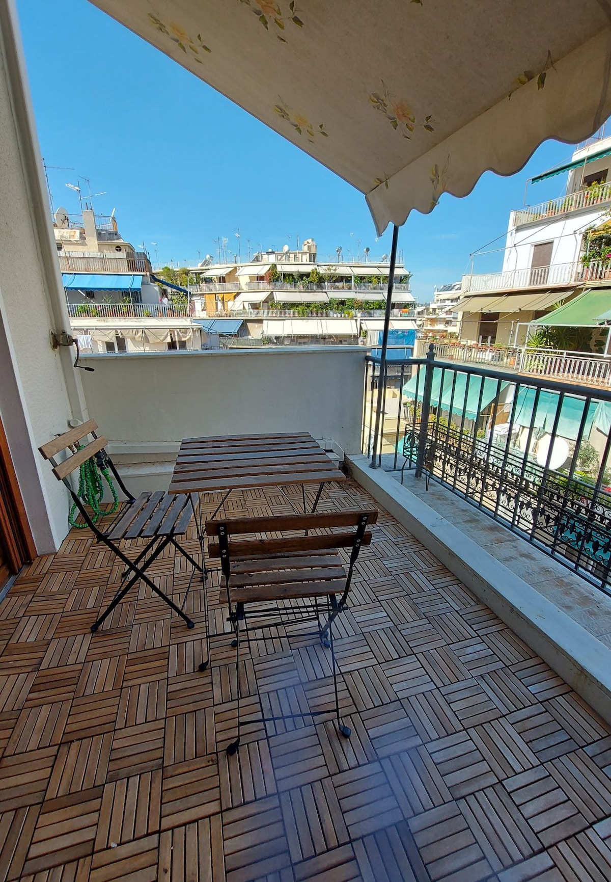 A small balcony is depicted, featuring a wooden table and two folding chairs. The area is surrounded by railings and overlooks neighboring buildings. A retractable awning provides shade, while the tiled flooring adds a touch of texture.