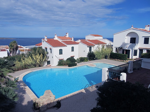 A clear view of a swimming pool is presented, bordered by lush greenery and white residential buildings with red roofs. The calm sea is visible in the background under a partly cloudy sky, suggesting a tranquil outdoor environment.