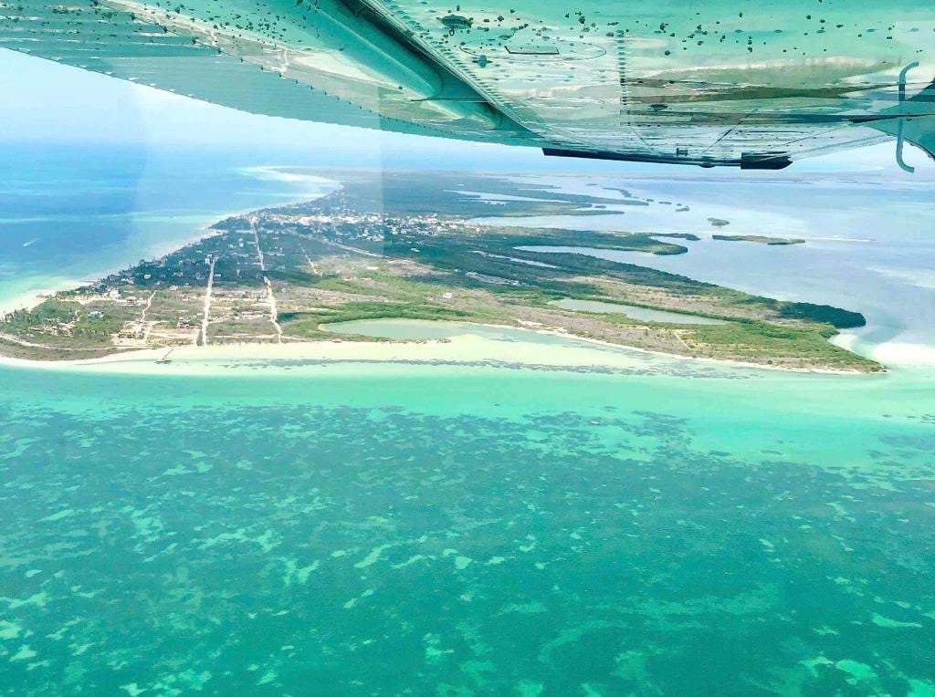 An aerial view of Holbox Island showcases lush green landscapes surrounded by clear turquoise waters. The coastline lines a developed area, with sandy beaches and patches of greenery visible among the natural environment, conveying the island's serene charm.