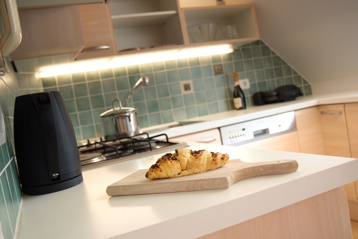A modern kitchen features light wood cabinetry and a turquoise tiled backsplash. A wooden cutting board displays a croissant with chocolate sprinkles. In the background, a kettle, saucepan, and a bottle of champagne are visible, highlighting a functional and well-equipped cooking space.
