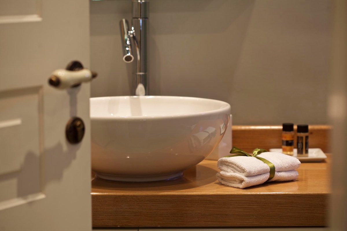 A modern bathroom is showcased, featuring a sleek, round sink on a wooden countertop. Neatly rolled towels are presented, accompanied by a small selection of toiletries. Light filters softly through an open door, adding warmth to the space.
